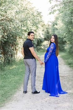 a man and woman holding hands while standing on a dirt road in front of trees