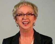 an older woman wearing glasses and a black blazer smiles at the camera while standing in front of a gray background