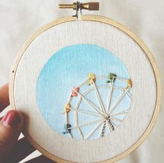 someone is holding up an embroidery project with ferris wheel in the middle and blue sky behind them