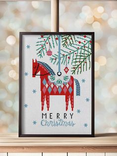 a cross stitch christmas card hanging on a window sill with lights in the background