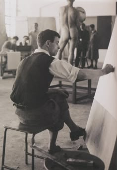 black and white photograph of a man sitting in front of an easel with his legs crossed