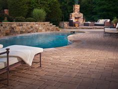 a brick patio next to a swimming pool with lounge chairs and an outdoor fireplace in the background