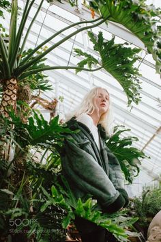 a blonde woman standing in a greenhouse surrounded by plants and greenery, looking up at the sky