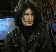 a man with long hair sitting in front of a tv screen wearing a leather jacket