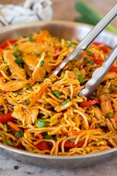 a close up of a bowl of food with noodles and chicken on the side next to two tongs