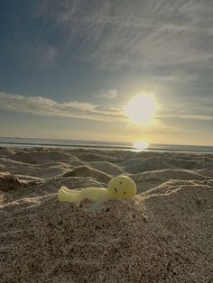 a yellow plastic toy laying in the sand at the beach with the sun behind it