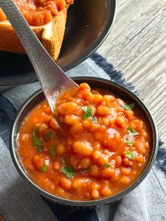 a spoon in a bowl filled with baked beans on top of a wooden table next to a piece of bread