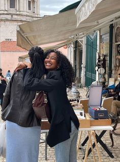 two women hugging each other in the middle of an open air market with people walking around