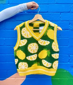 a sweater with lemons on it hangs against a blue brick wall in front of a person's hand