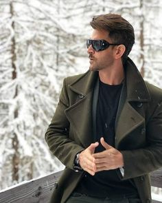 a man wearing sunglasses standing in front of snow covered trees and holding his hands together