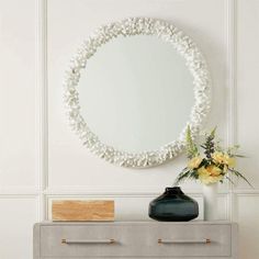 a round mirror hanging on the wall above a dresser with flowers and a vase in front of it