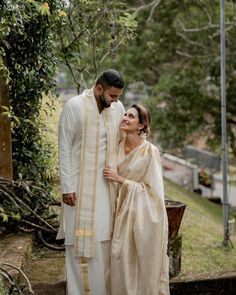 a man and woman standing next to each other in front of some trees with their arms around each other