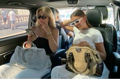 two women sitting in the back seat of a car looking at their cell phones and purses