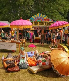 people are sitting on the grass under umbrellas at an outdoor market with tents in the background