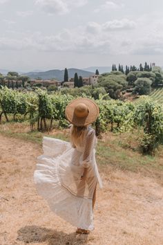 a woman in a white dress and straw hat walks through the vineyards with her back to the camera