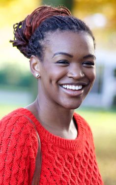 a smiling woman in an orange sweater with braids and a brown bag on her shoulder