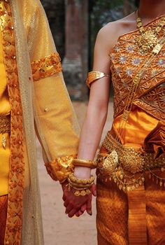 two people holding hands while dressed in gold and orange outfits with elaborate jewelry on their arms