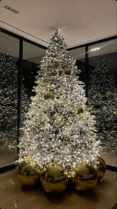 a white and gold christmas tree in a room with large windows, decorated with shiny balls