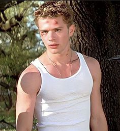 a young man standing in front of a tree wearing a white shirt and silver necklace