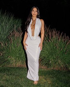 a woman wearing a white dress standing in front of some tall grass and bushes at night