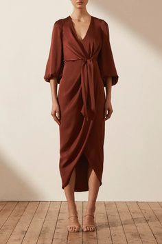 a woman in a brown dress stands on a wooden floor with her hands behind her back