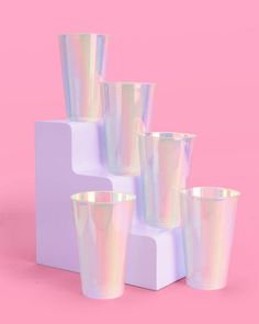 a set of four iridescent cups sitting on top of a white block against a pink background