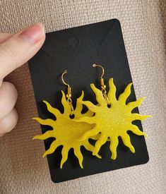 a pair of yellow sun earrings sitting on top of a piece of black paper next to a hand