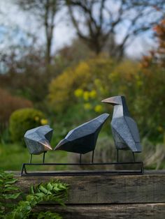 two metal birds sitting on top of a piece of wood next to plants and trees