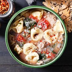 a green bowl filled with pasta and meat soup on top of a wooden table next to bread