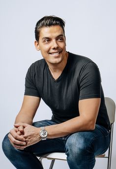 a man sitting on top of a white chair wearing a black shirt and blue jeans