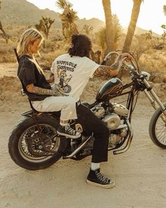 a man and woman sitting on a motorcycle in the desert with palm trees behind them