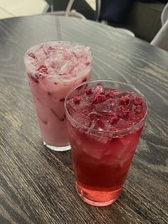 two drinks sitting on top of a wooden table next to each other, one with ice and the other with raspberries