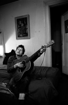 a man sitting on top of a bed holding a guitar in his right hand and looking up at the ceiling