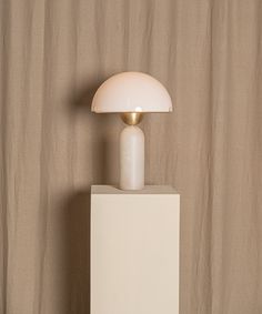 a white table lamp sitting on top of a white pedestal next to a brown curtain