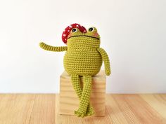 a crocheted frog sitting on top of a wooden block wearing a red hat