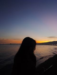 a woman standing on top of a beach next to the ocean at sunset with her hair blowing in the wind