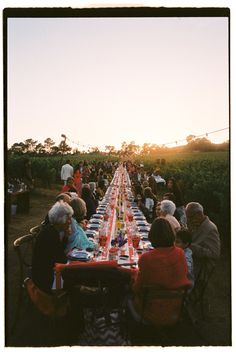 a long table with many people sitting at it in the middle of a field during sunset