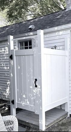 a white outhouse sitting in the middle of a yard next to a tree and wicker chair