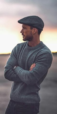 a man with his arms crossed standing in front of the sun wearing a hat and sweater