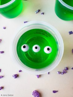 two green cups with googly eyes and purple confetti scattered around them on a white surface