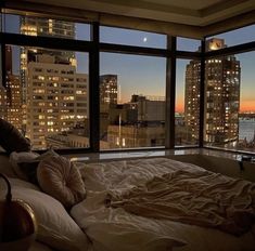 an unmade bed in front of a large window overlooking the city at night time