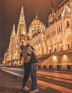 two people standing on the sidewalk in front of a large building with steeples at night