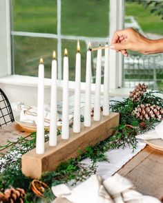 a person lighting candles on a table with pine cones and greenery in the foreground