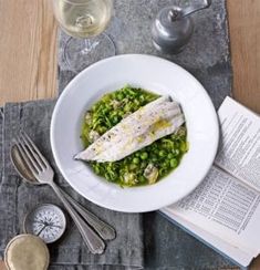 a white plate topped with fish and peas next to a glass of wine on top of a table