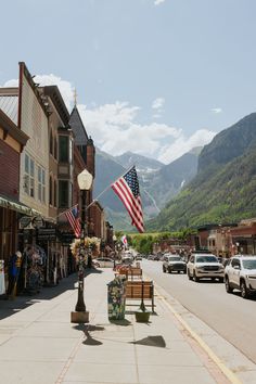 an american flag flying in the wind on a street with buildings and mountains behind it