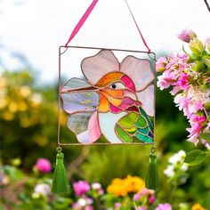 a stained glass humming bird hanging from a pink flowered plant with green tassels