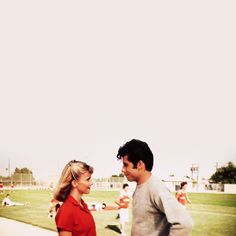 a man and woman standing next to each other in front of a baseball field on a sunny day