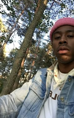 a young man wearing ear buds and a denim jacket is standing in front of trees