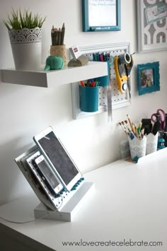 a white desk topped with lots of office supplies next to a wall filled with pictures