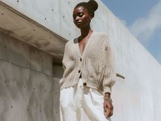 a woman standing in front of a concrete wall wearing white pants and a cardigan sweater